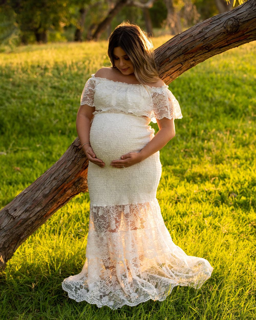 A pregnant woman in a white lace maternity gown poses in golden sunlight near a fallen tree trunk in a grassy field.