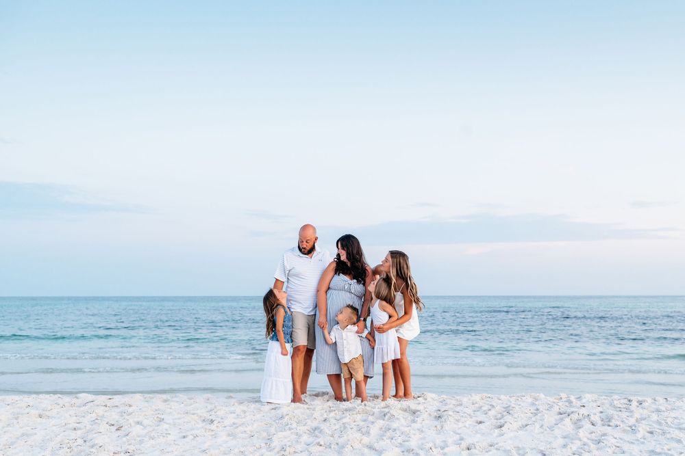 People wearing white outfits walk along a sunny beach with turquoise ocean waves and clear blue sky in background.
