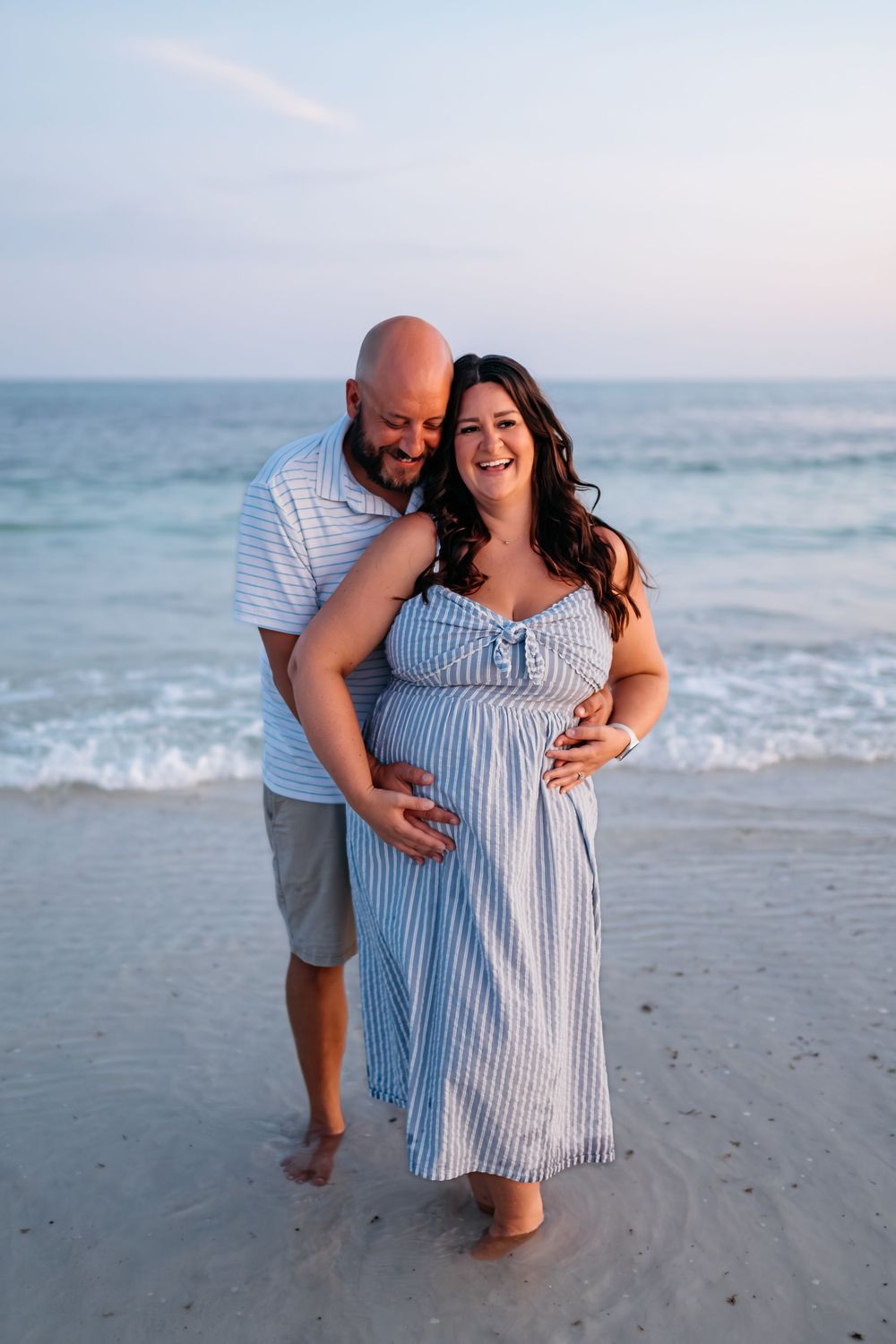 A couple shares a tender moment on the beach at sunset in matching light blue and white outfits.