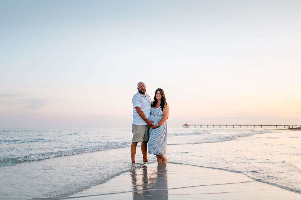 A couple stands together on a beach at sunset with a pier visible in the distance.