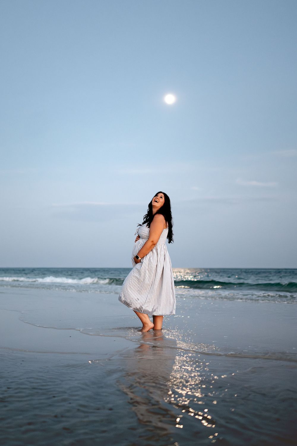 A woman in a white flowing dress poses gracefully in shallow ocean waters at sunset creating ethereal reflections.
