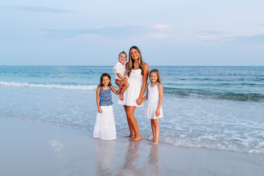 Family wearing coordinated white and blue outfits poses together on a beach at dusk with ocean waves in the background.