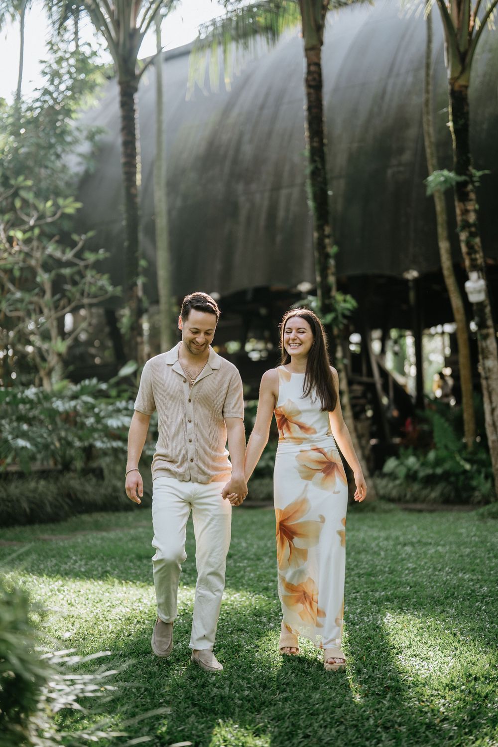 A couple strolls together on a sunlit lawn surrounded by palm trees and tropical foliage.