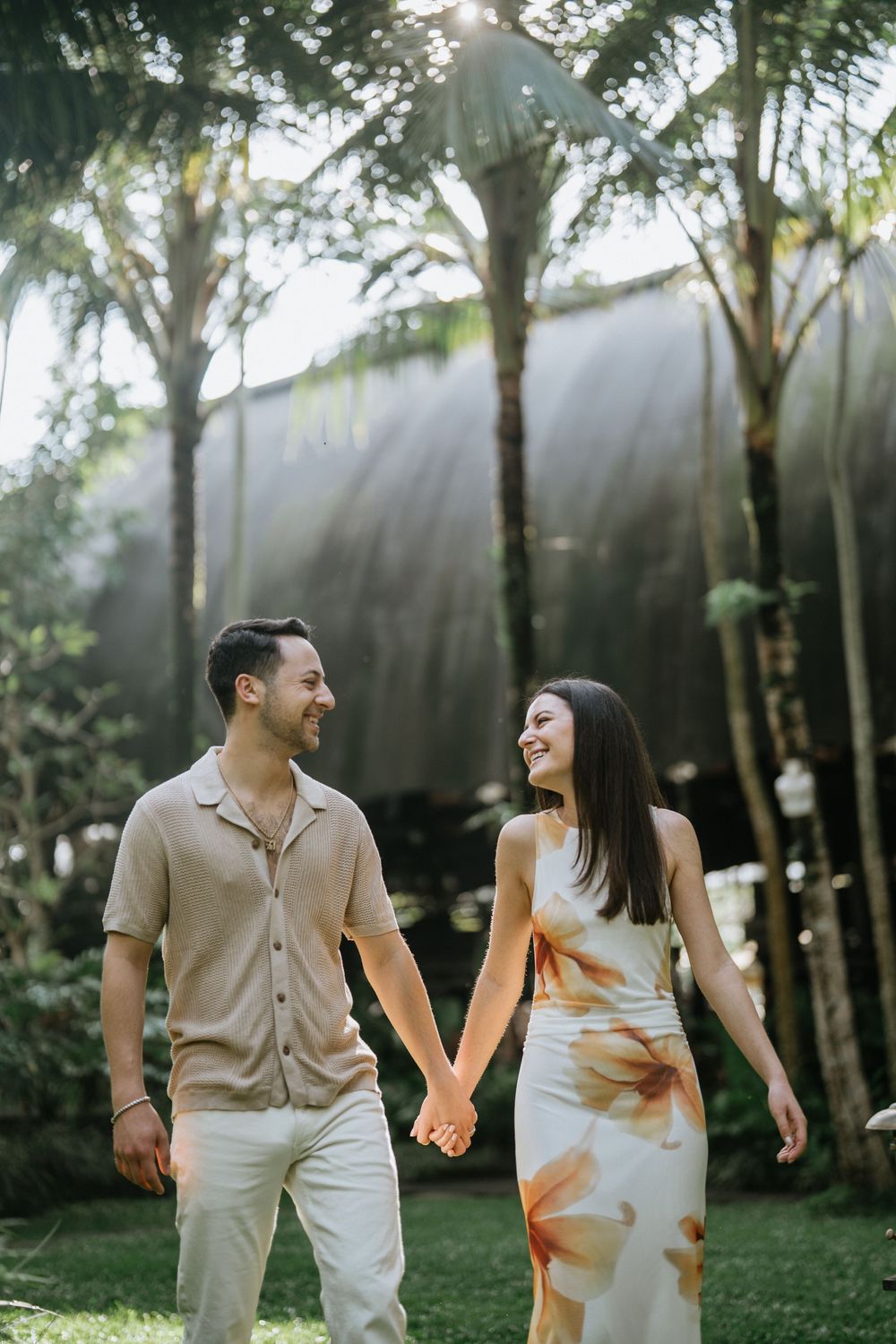A playful couple in coordinated outfits dances on a grassy lawn with palm trees in the background.