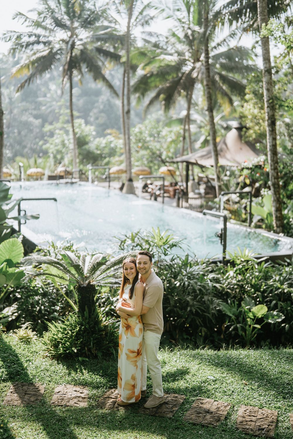 Couple embraces in a tropical resort setting with palm trees and an infinity pool in the background.