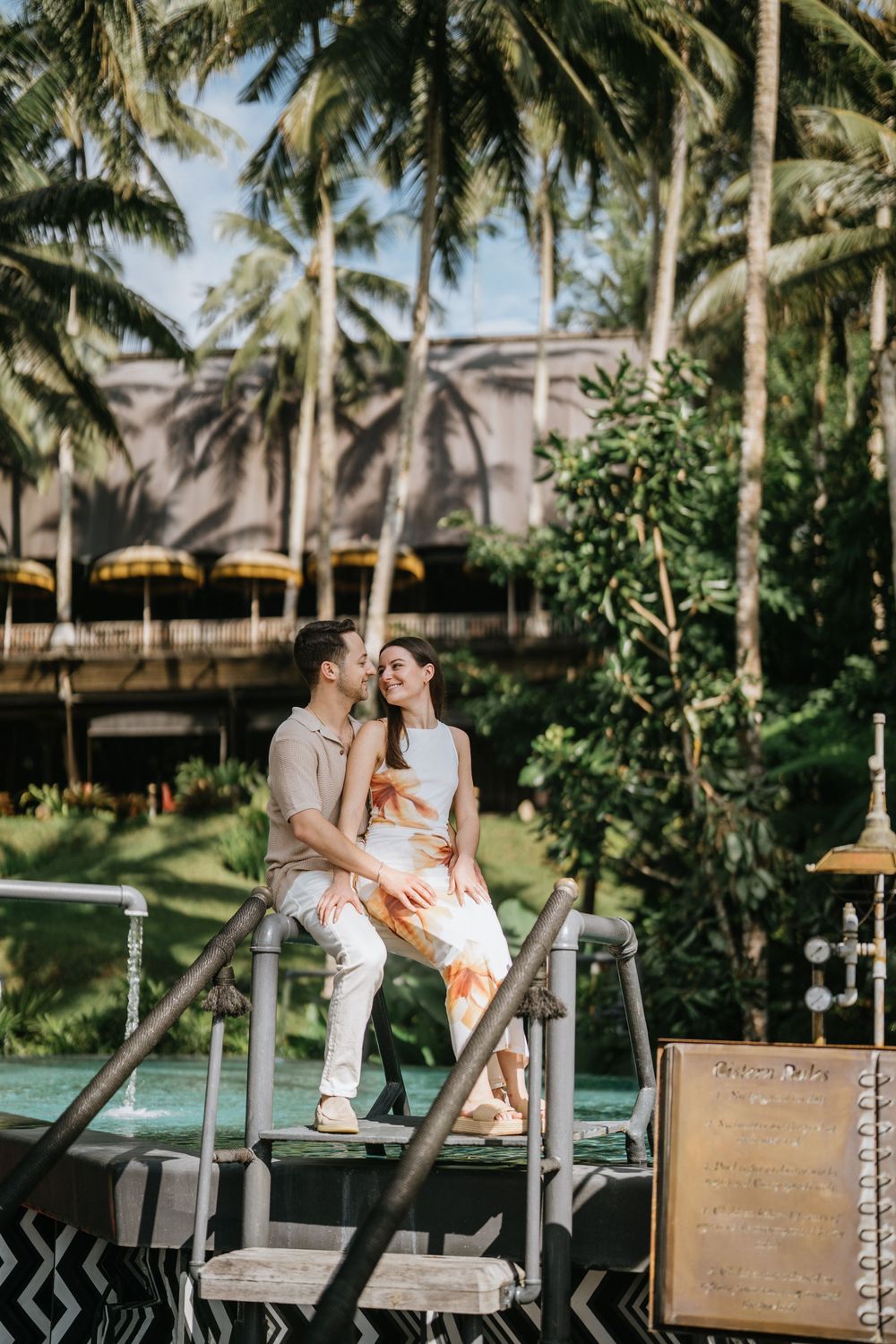 A couple shares a romantic moment while sitting on a vintage wooden cart surrounded by palm trees.