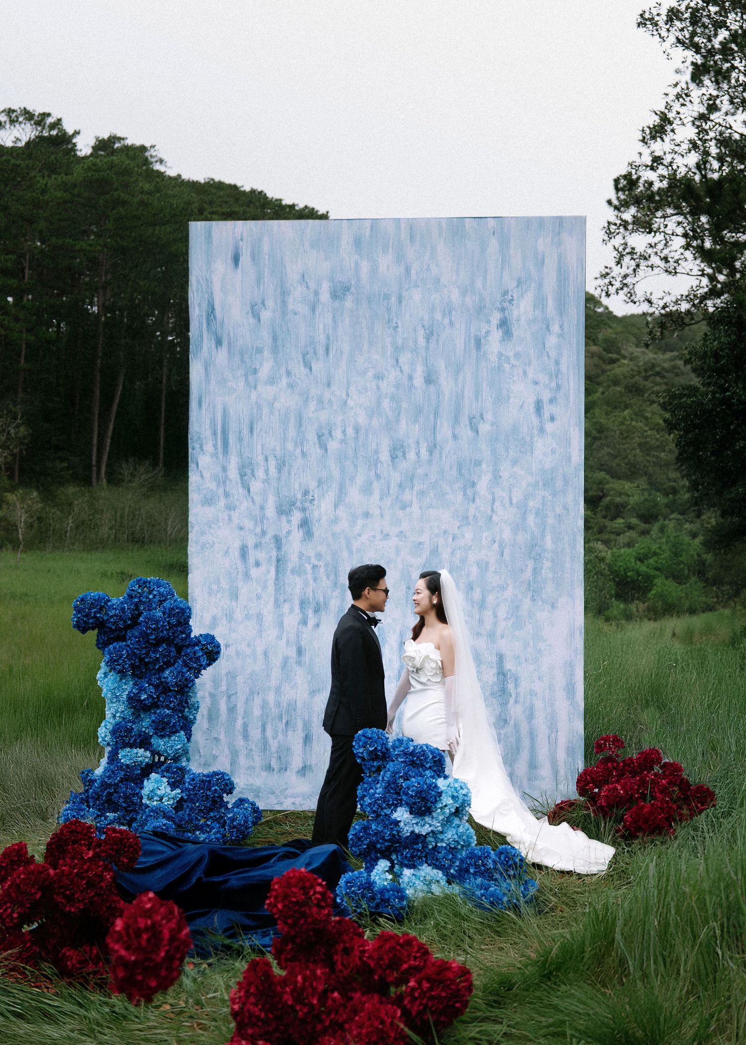 Bride and groom stand before a large blue abstract backdrop surrounded by blue hydrangeas and red peonies outdoors.