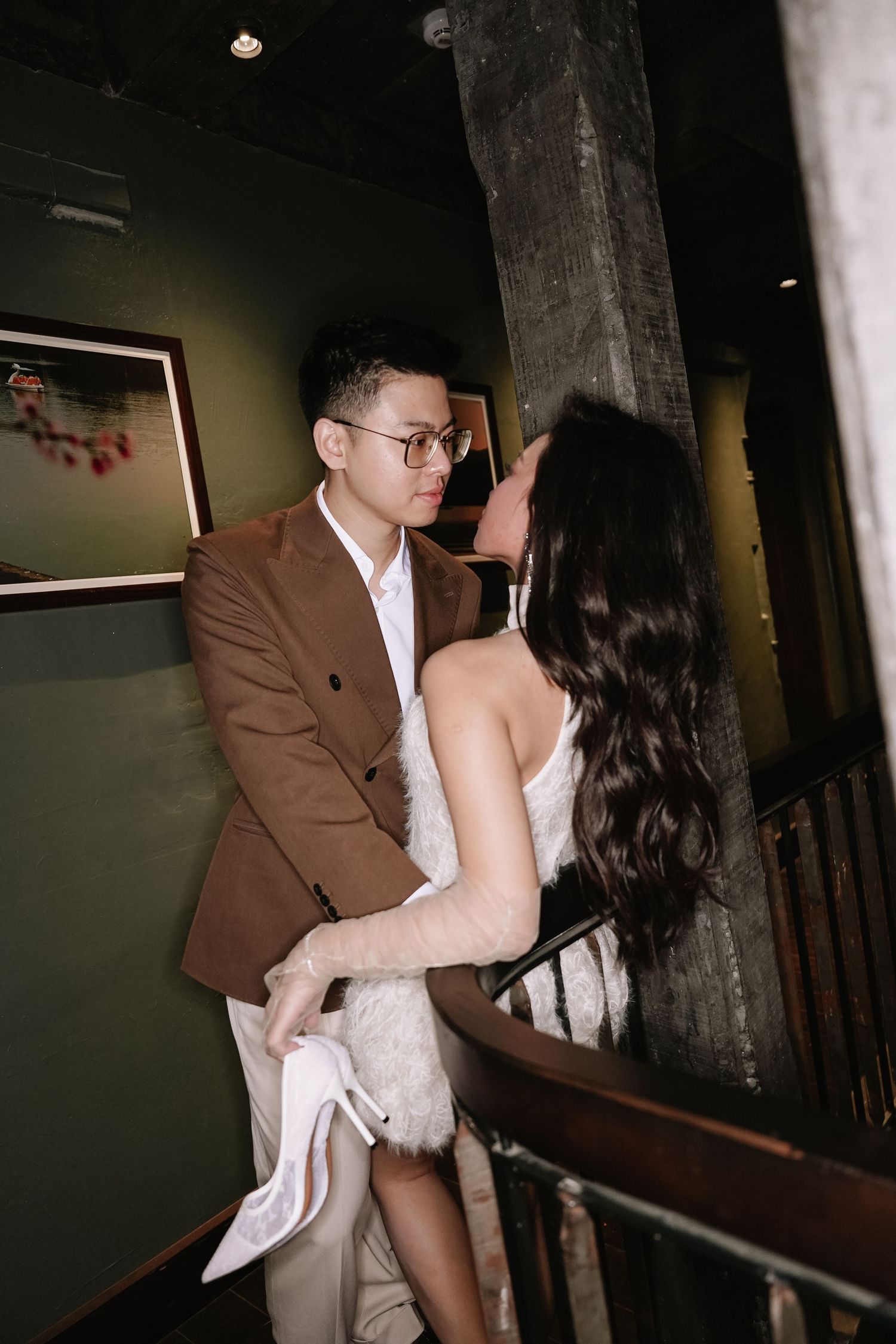 Romantic couple gazing at each other on a staircase, woman holding white heels in dim lighting.