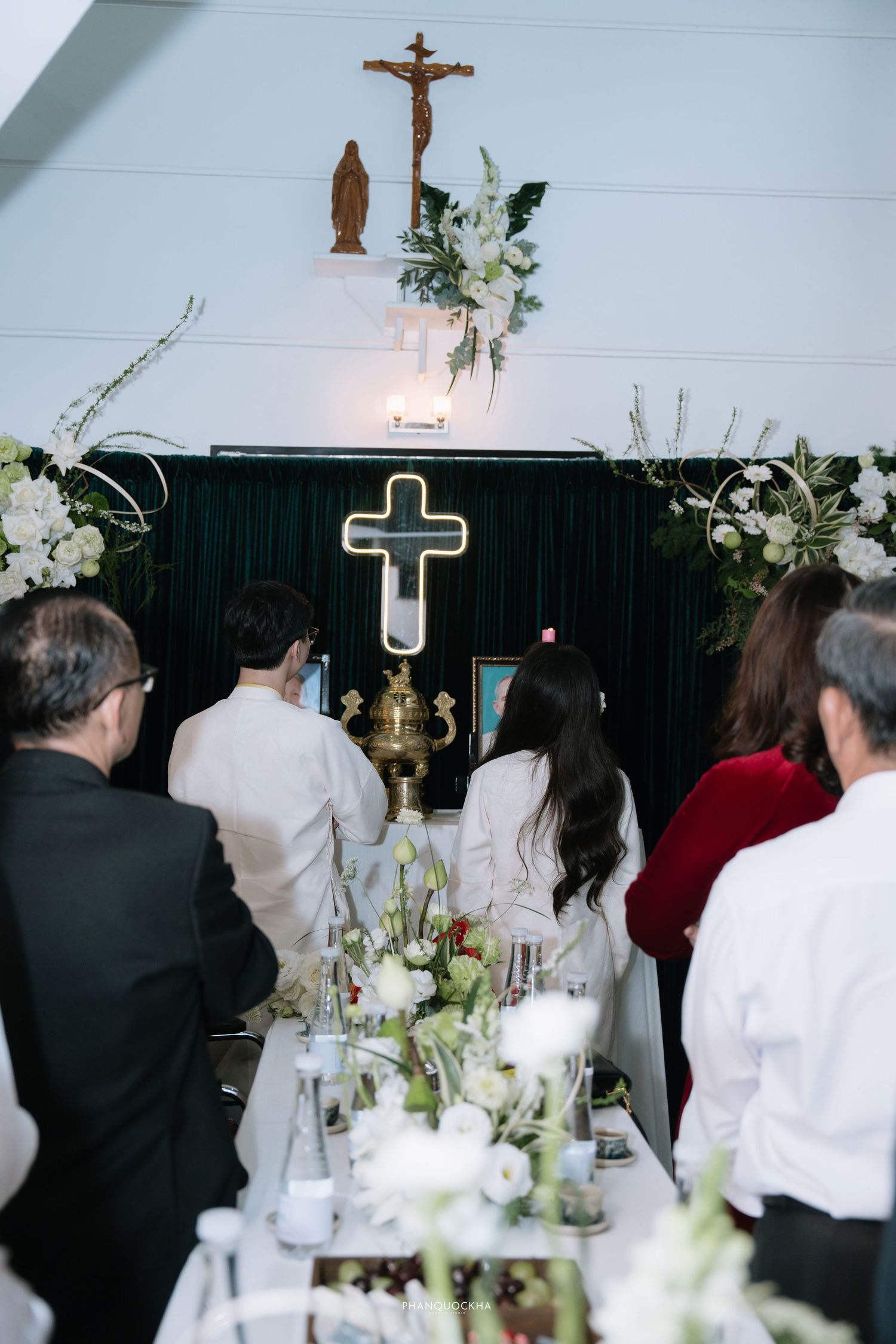 Guests in white attire pray before a Christian cross altar adorned with white floral arrangements.