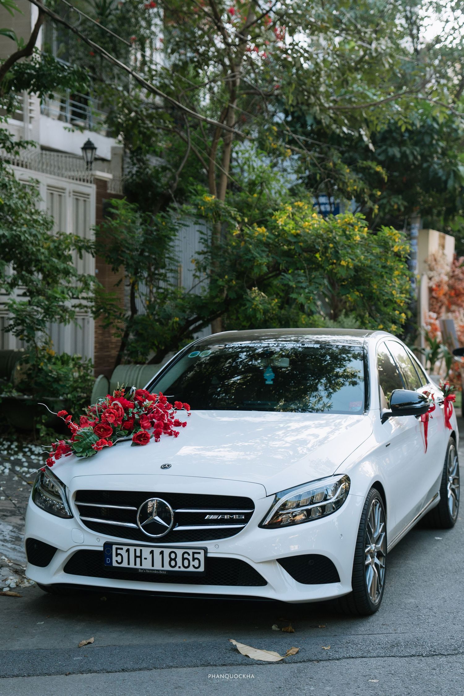 White Mercedes-Benz C-Class AMG wedding car decorated with red flowers on a tree-lined street.
