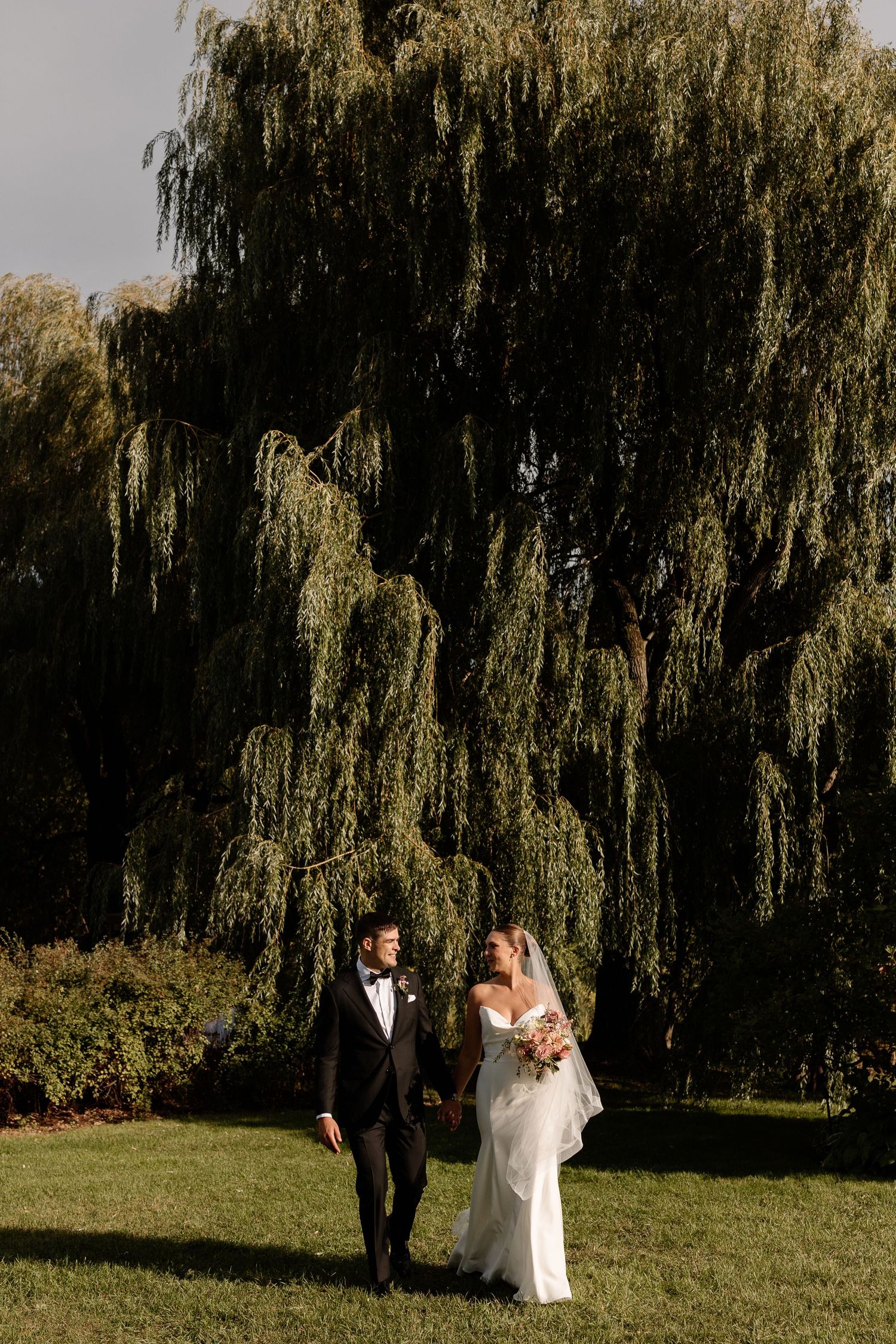 A wedding couple shares intimate moments in a garden setting with weeping willow trees. Aliza Dworkind Montreal wedding photographer.