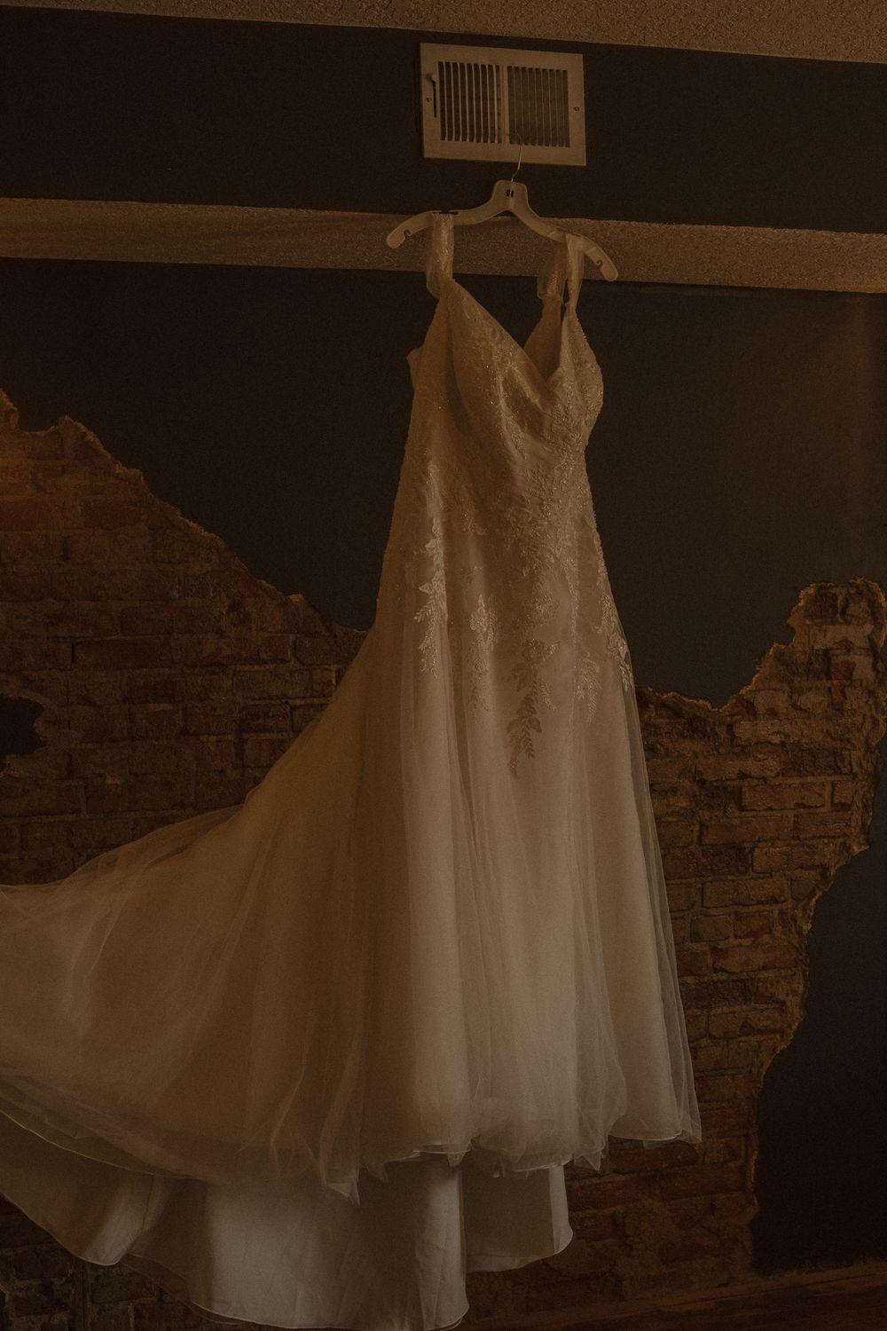 A delicate white lace wedding gown displayed against a rustic brick wall.