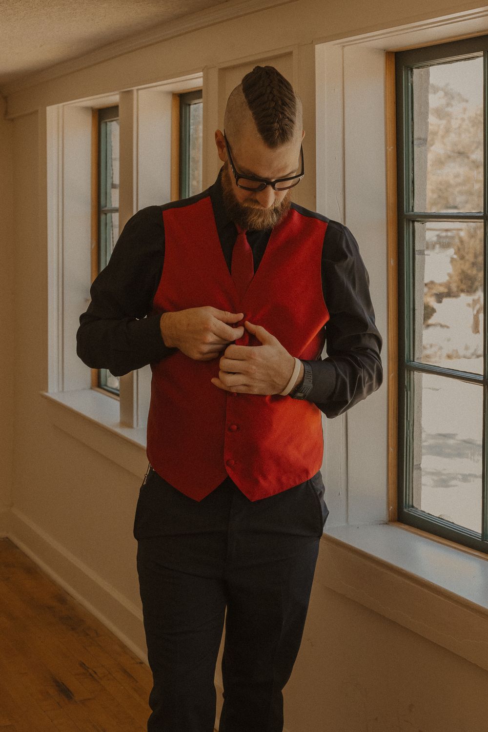 Person wearing a black suit and red vest adjusts their attire near windows.