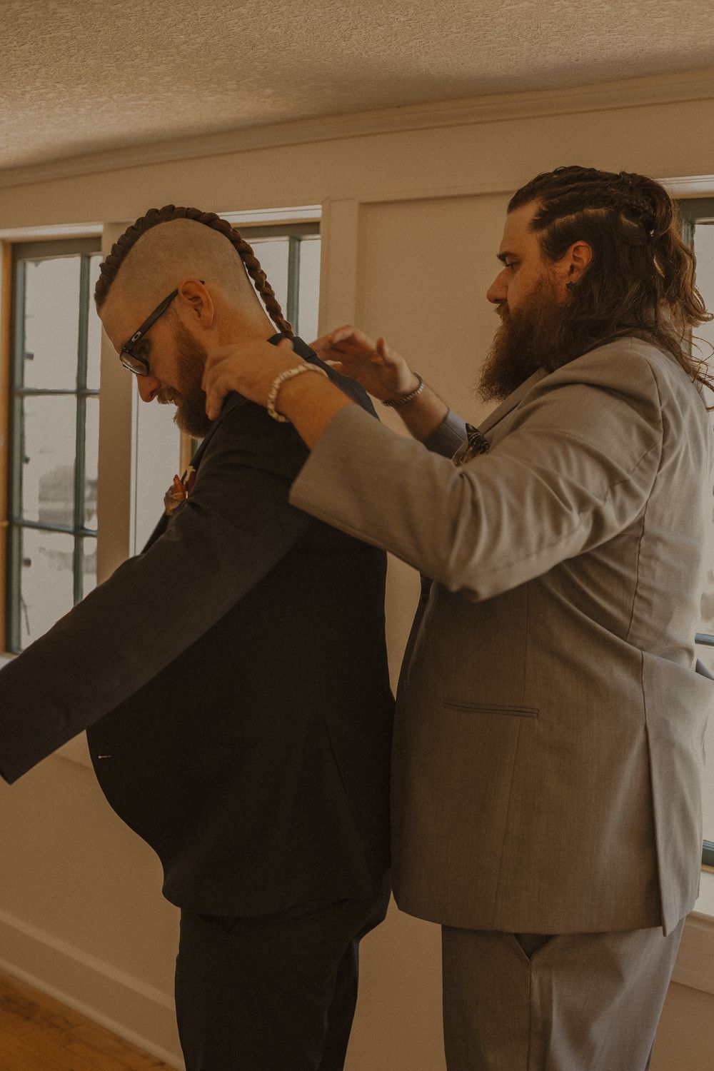Two people in formal attire getting ready in a warmly lit room with windows.