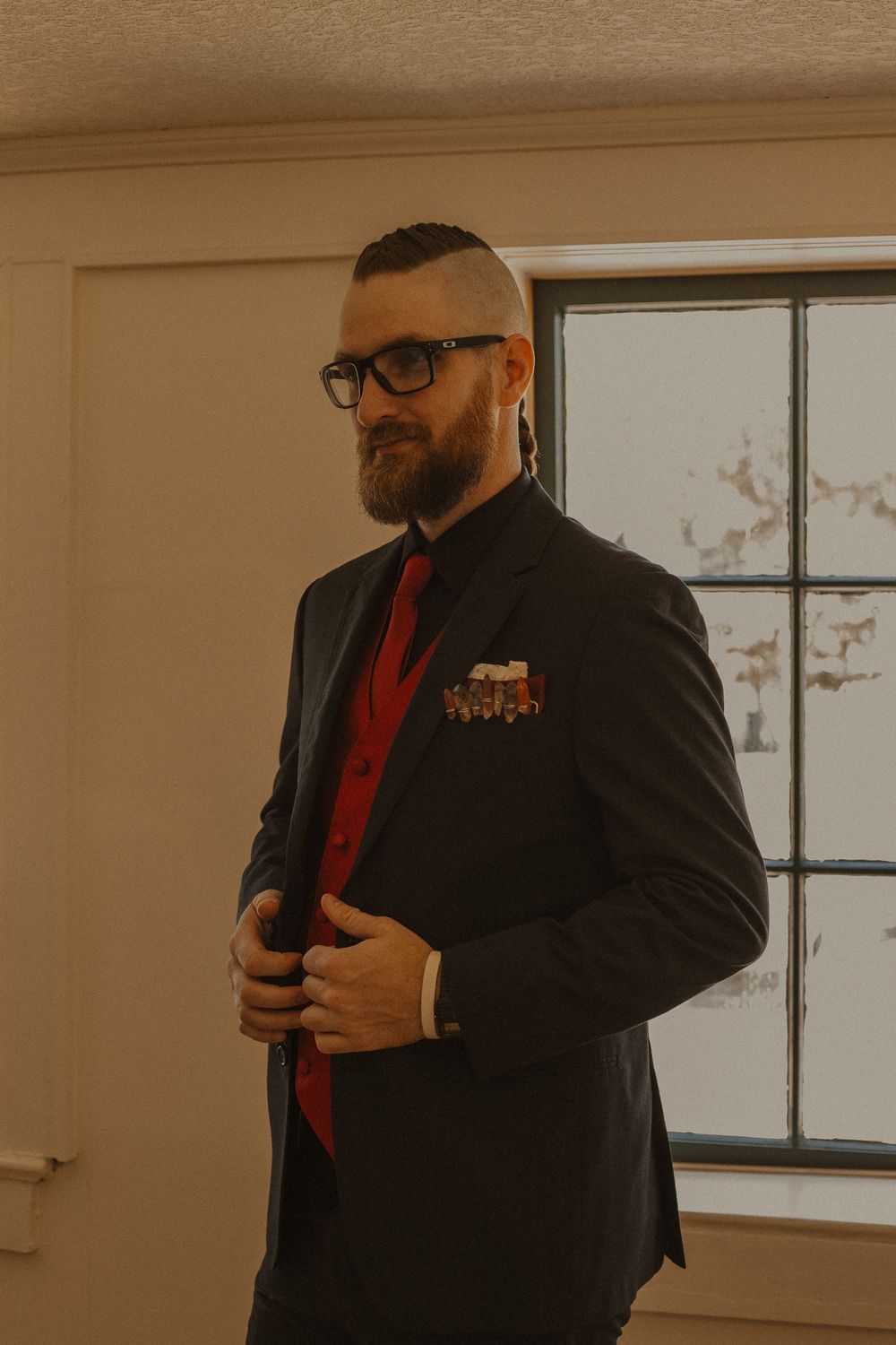 Figure in formal black suit with red vest and glasses stands in sunlit room.