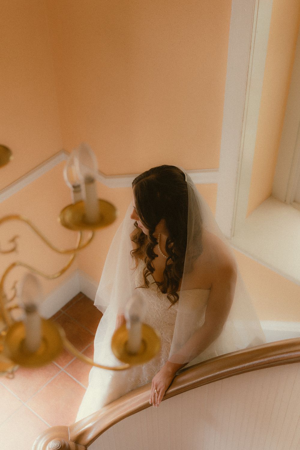 Bride in white gown and veil descends elegant staircase in warm golden light.