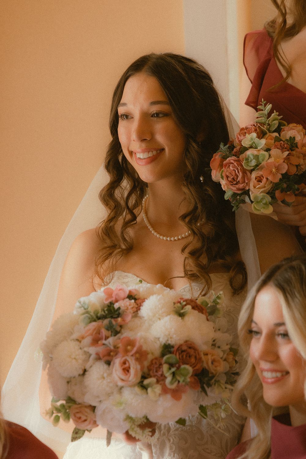 Smiling bride in strapless gown holds white and pink bouquet, wearing pearl necklace and veil.