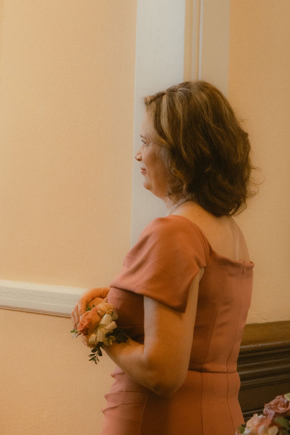 A woman in a dusty rose dress holds a small floral bouquet while standing profile against a warm beige wall.