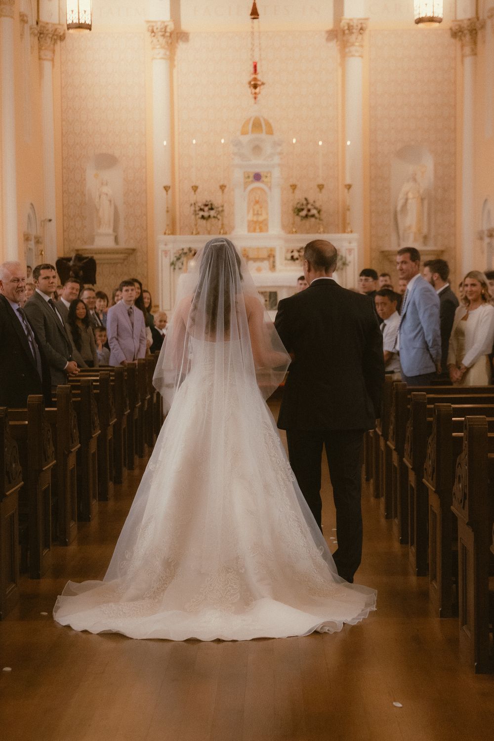Bride in white gown and veil walks down church aisle with escort toward altar during wedding ceremony.