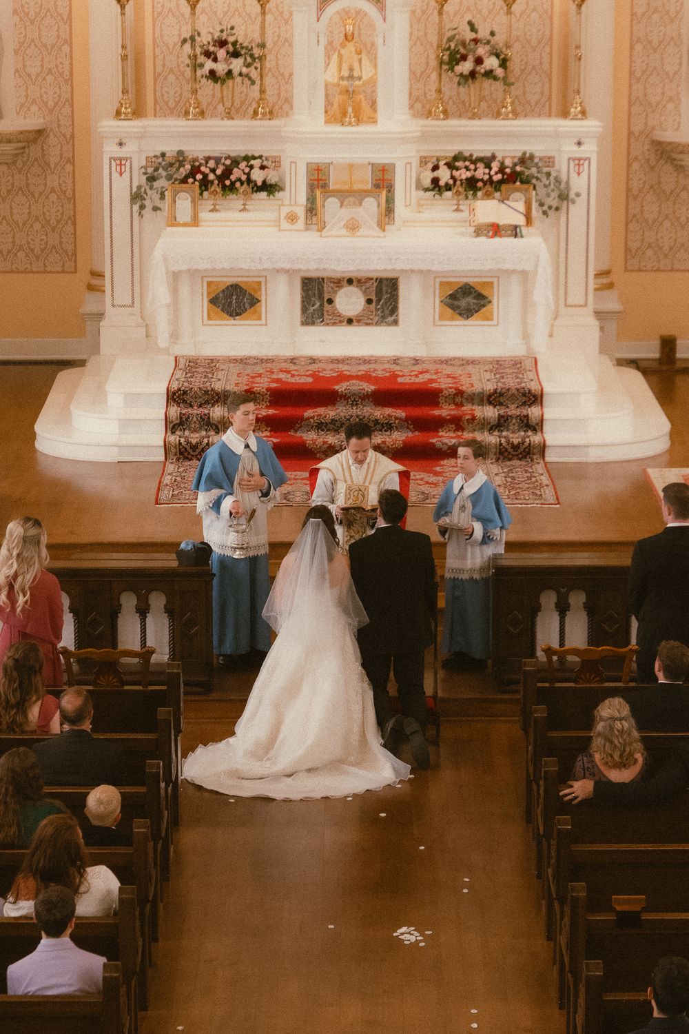 Bride and groom exchange vows at ornate church altar with clergy in blue robes and floral arrangements during wedding ceremony.