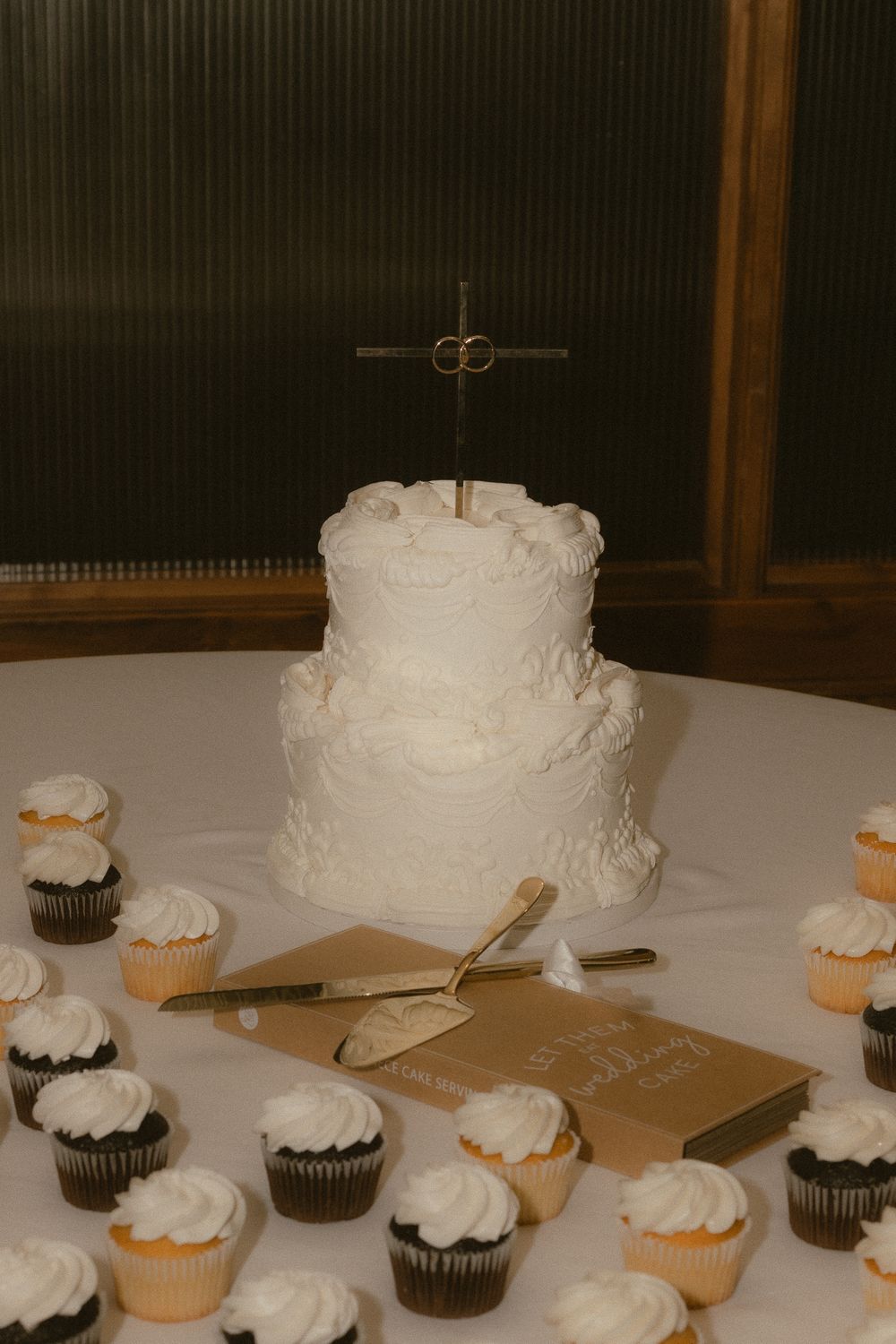 Two-tier white wedding cake with cross topper surrounded by chocolate and vanilla cupcakes on a white tablecloth.