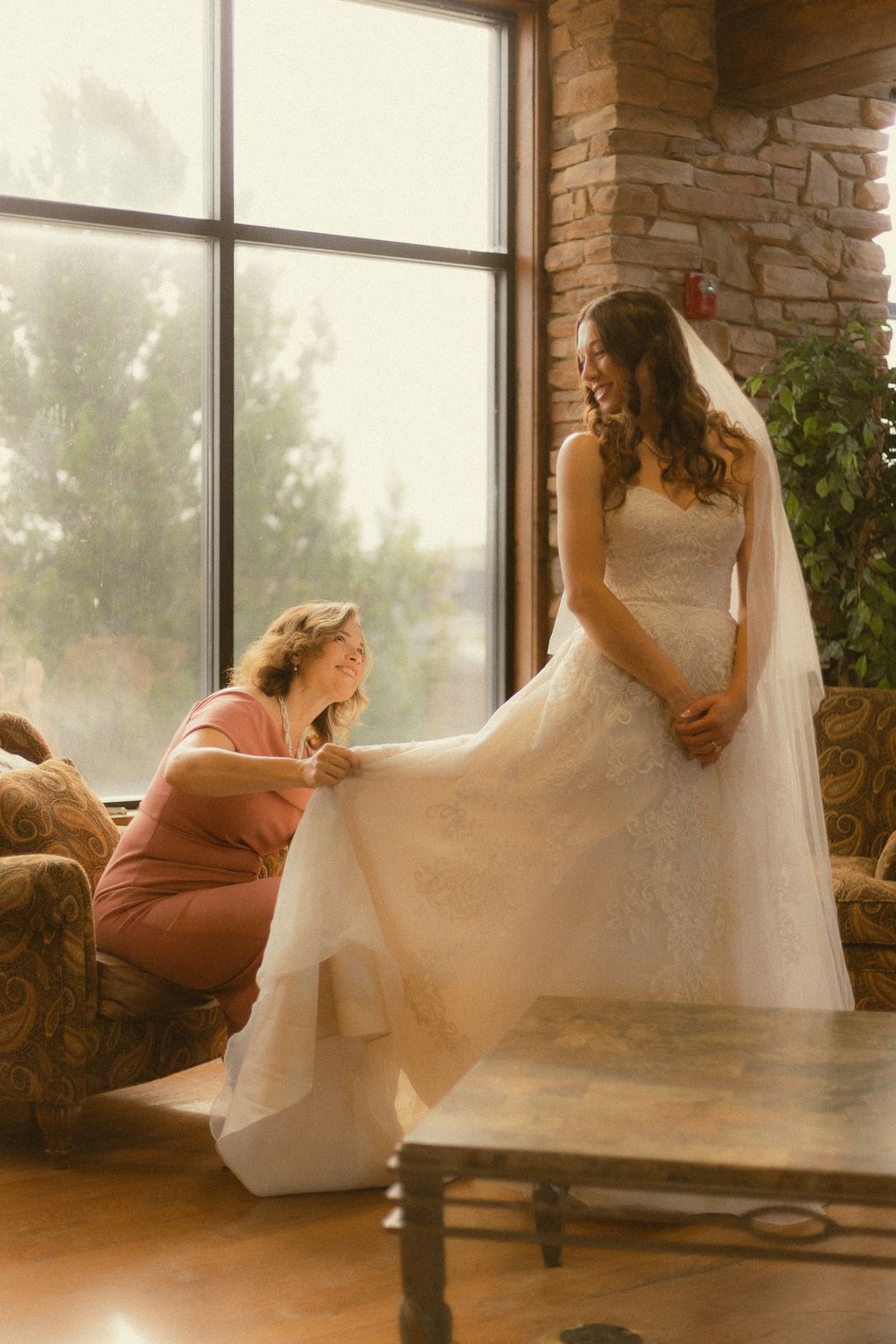 A bride in a white lace gown and veil stands as a woman in pink adjusts her dress near a large window.