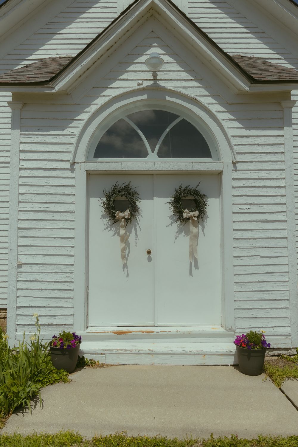 White country church entrance with double doors adorned with greenery wreaths, arched window above, and flower pots on steps.