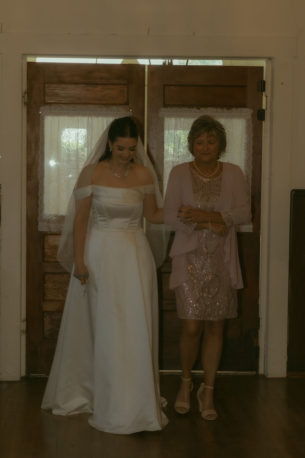Bride in white gown and veil stands with older woman in pink dress by wooden doors at wedding venue.