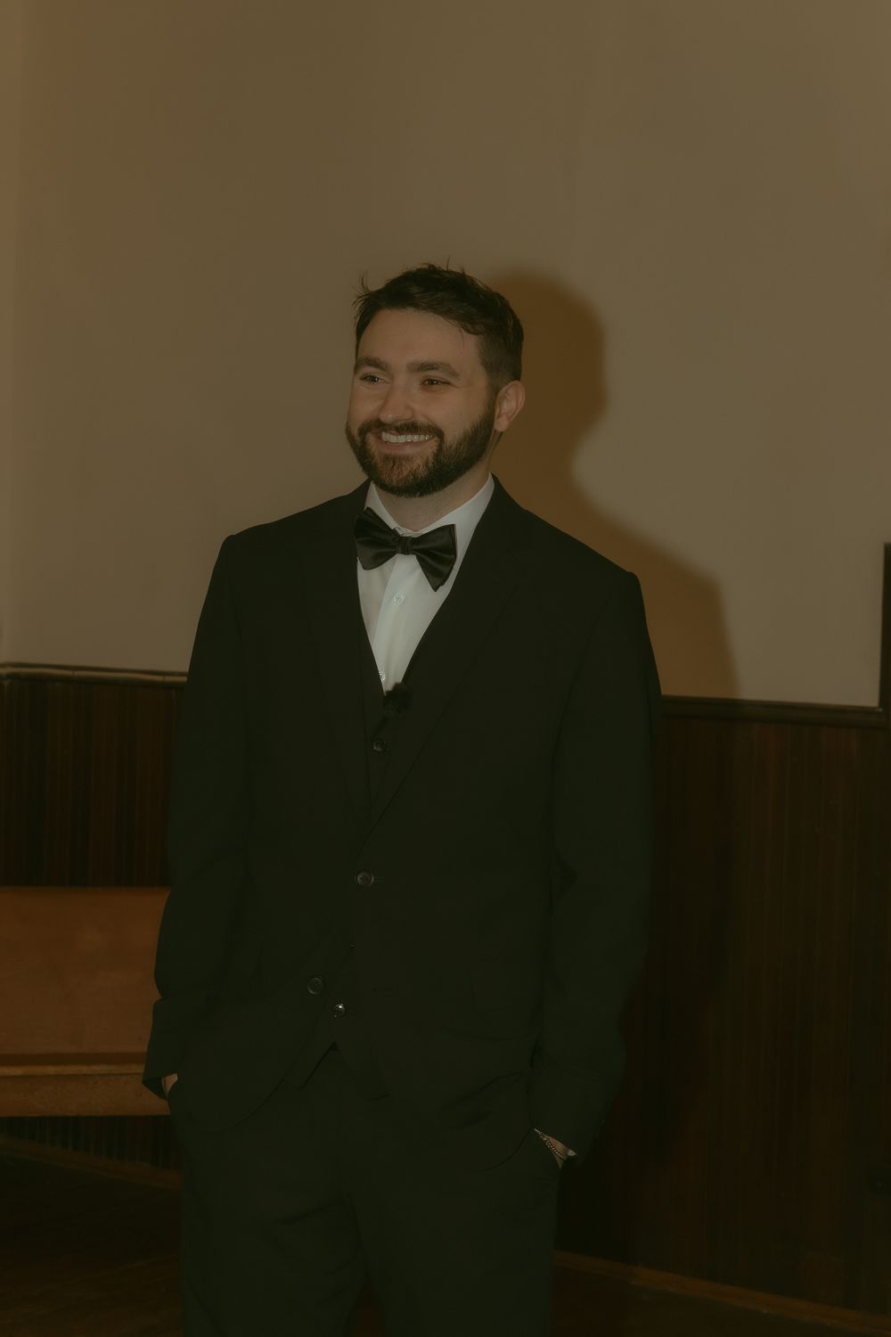 A smiling man with a beard wearing a black tuxedo with bow tie stands in a warmly lit room.