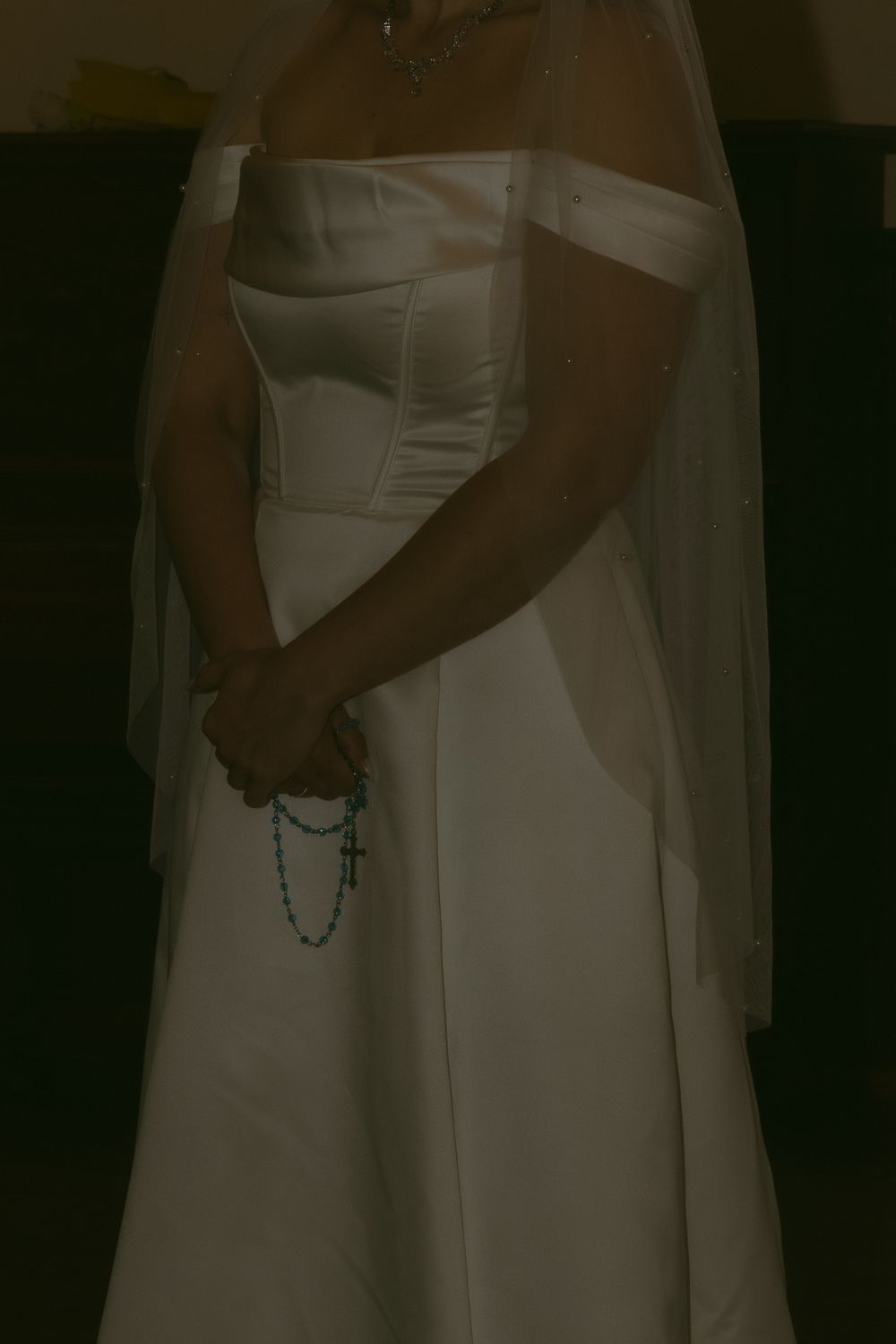 Bride in white strapless wedding gown holding blue rosary beads with cross, wearing veil, standing in dimly lit room.