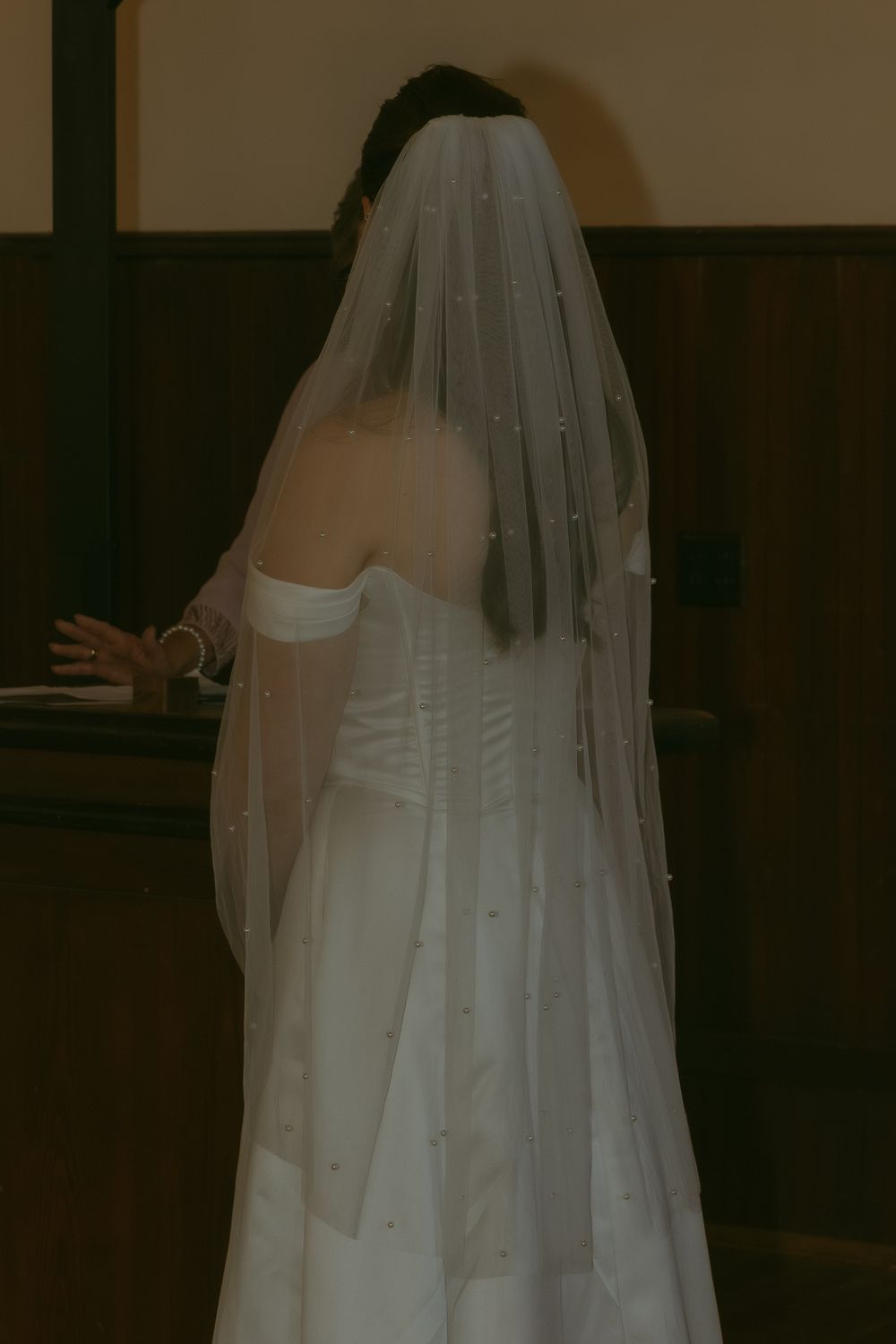 Bride in white strapless wedding gown with pearl-trimmed veil, viewed from behind in a church.