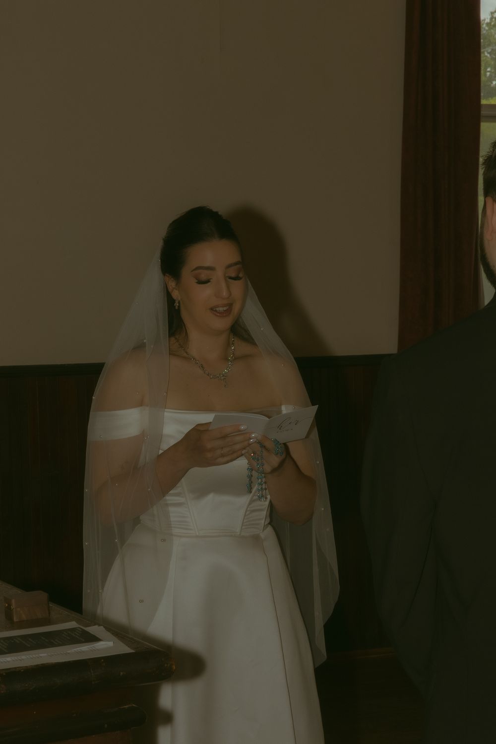 A bride in a white off-shoulder gown and veil reads her vows during an indoor wedding ceremony.
