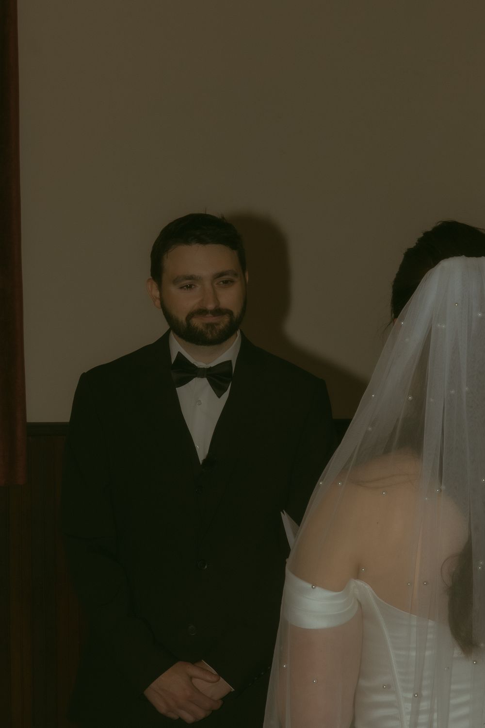 Groom in black tuxedo and bow tie smiling at bride in white strapless wedding dress with veil during ceremony.