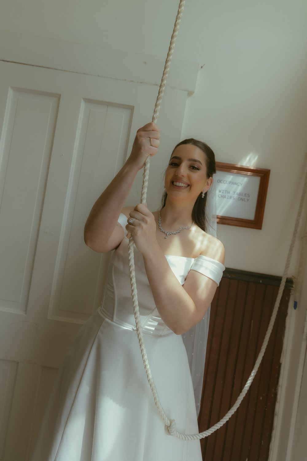 Smiling bride in white off-shoulder gown holding a rope bell pull inside a church.