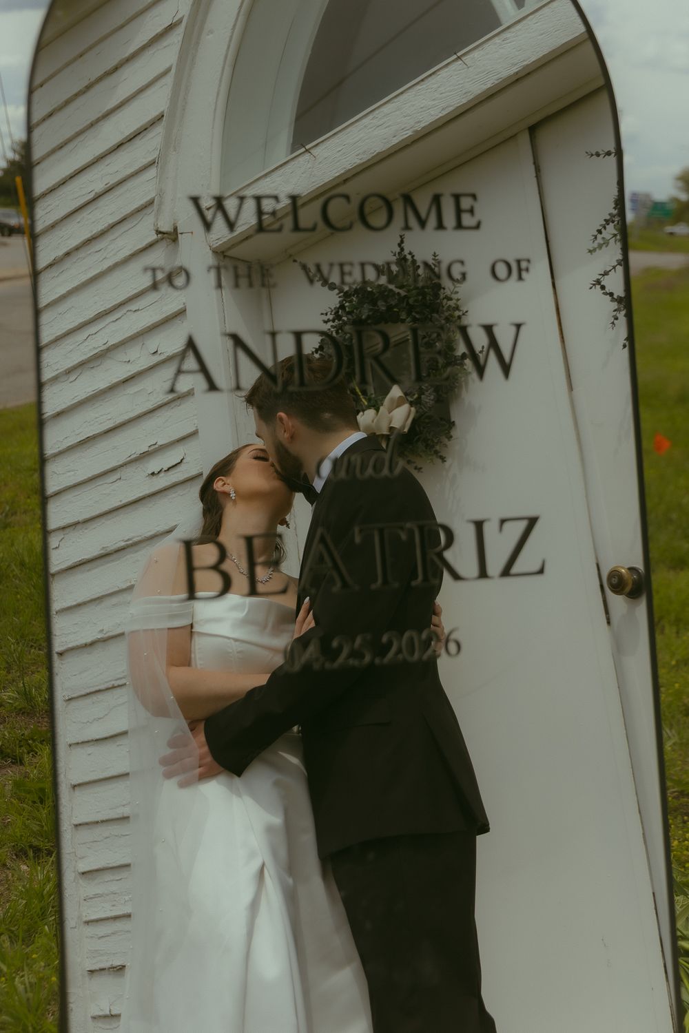 Bride and groom share a romantic kiss in front of a wedding welcome sign for Andrew and Beatriz on 10.25.2026.