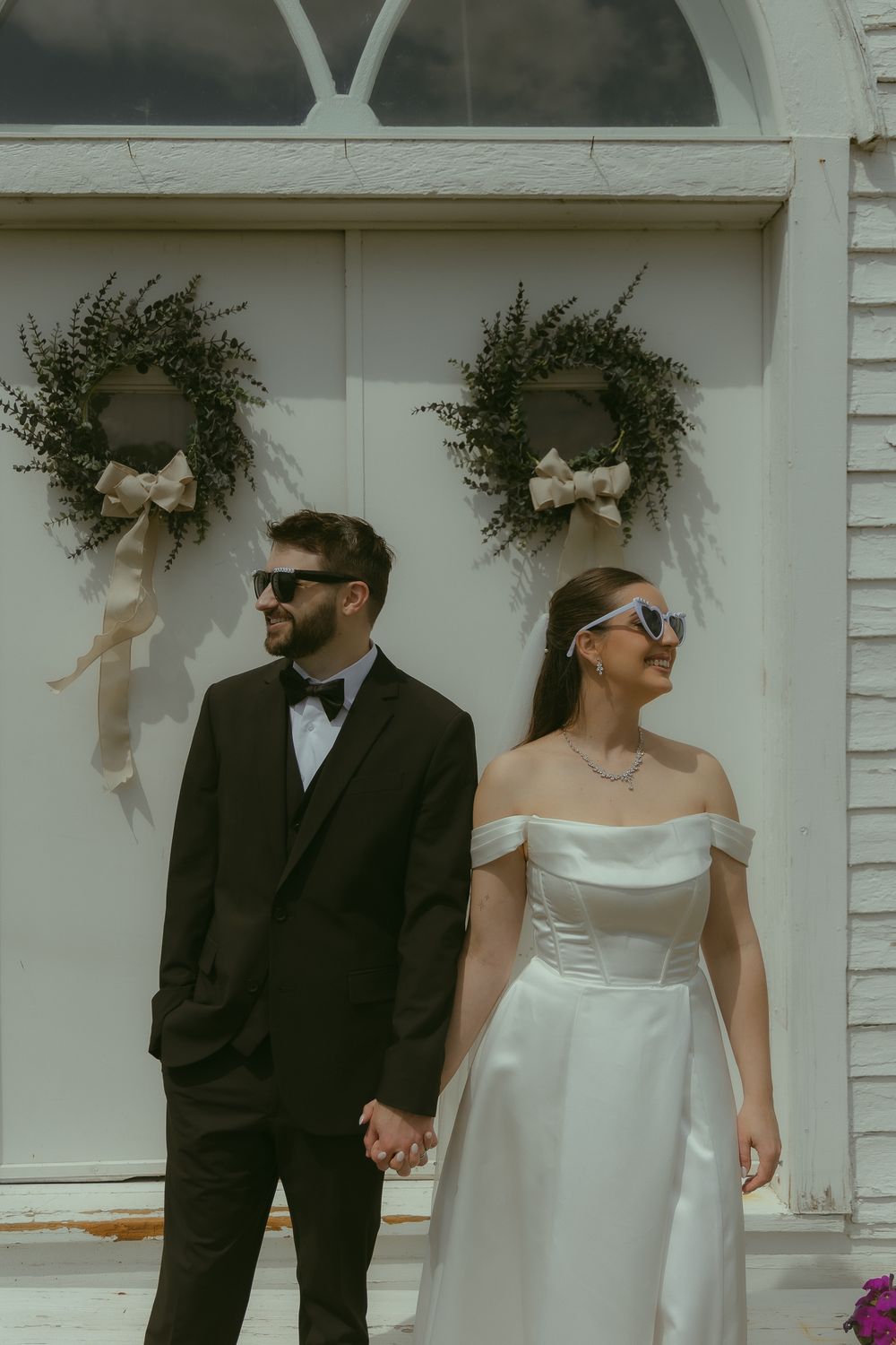 A bride and groom in sunglasses hold hands before a white chapel door adorned with wreaths on their wedding day.