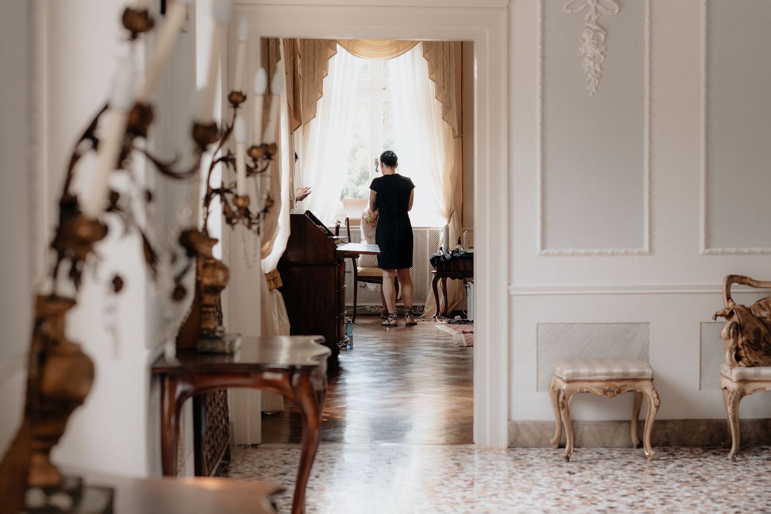 A silhouetted figure stands at a bright window in an elegant room with classical decor.