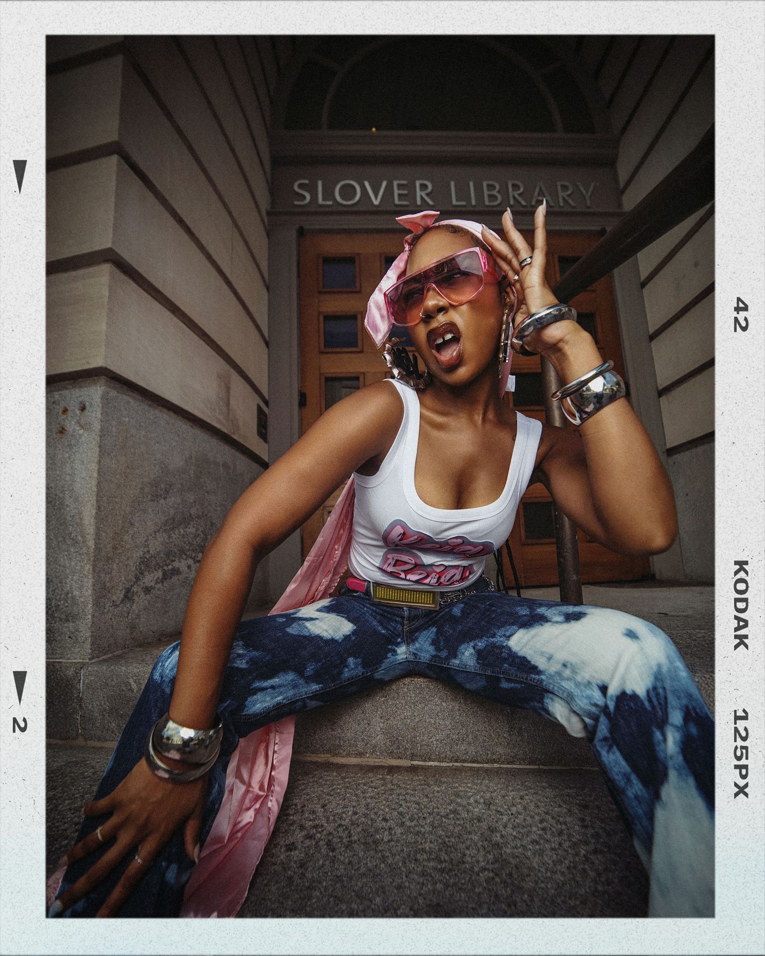 A person in tie-dye jeans and a tank top poses energetically on steps outside Slover Library.