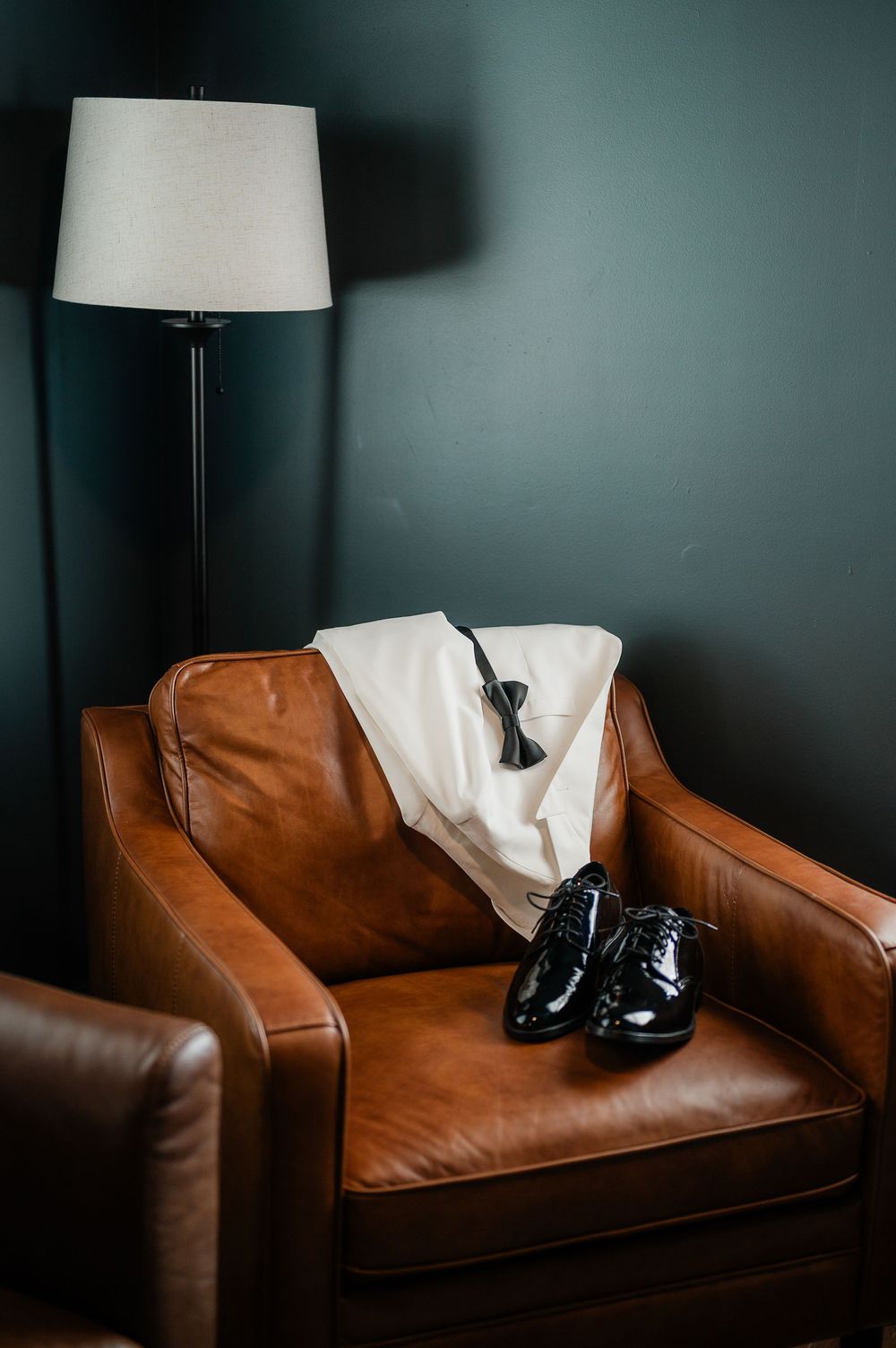 Black formal shoes rest on a brown leather couch against a dark wall with a lamp.