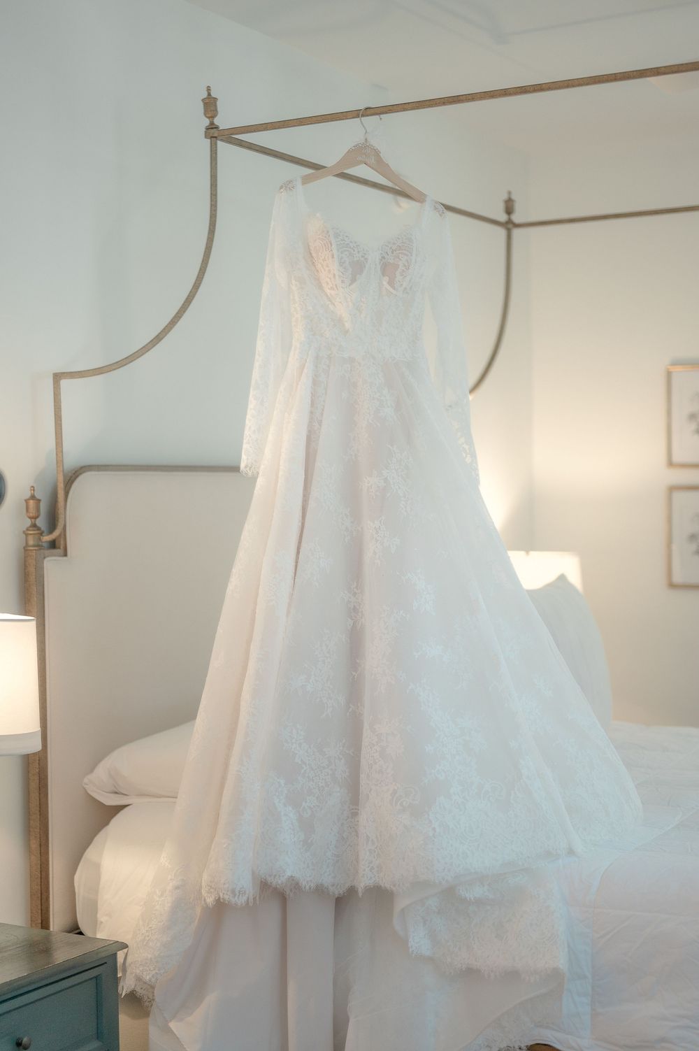 A delicate white lace wedding dress hangs from an elegant canopy bed frame in a softly lit bedroom.