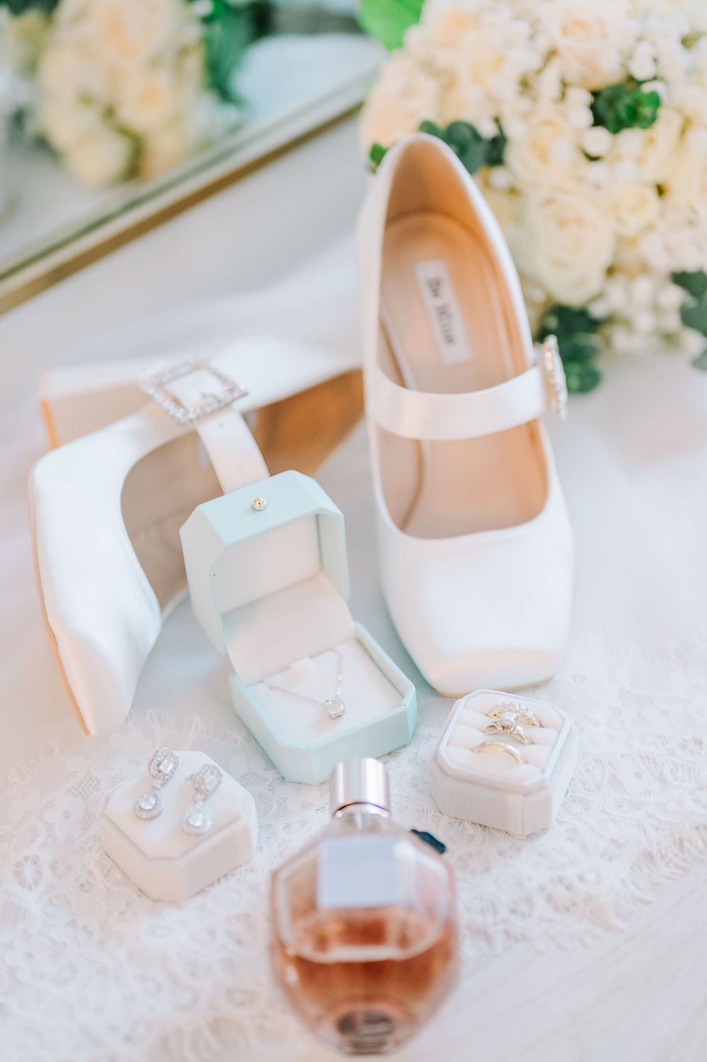 Elegant white baby shoes and jewelry displayed on delicate lace fabric with soft floral accents in the background.
