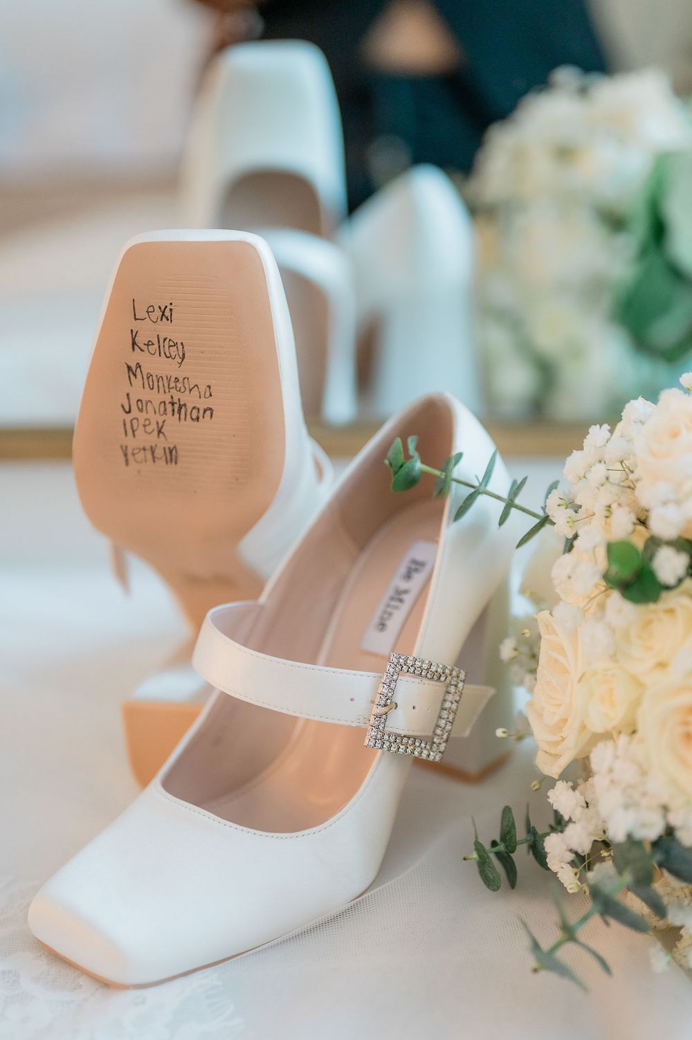 White bridal heels with handwritten messages on the soles displayed next to delicate white floral arrangements.