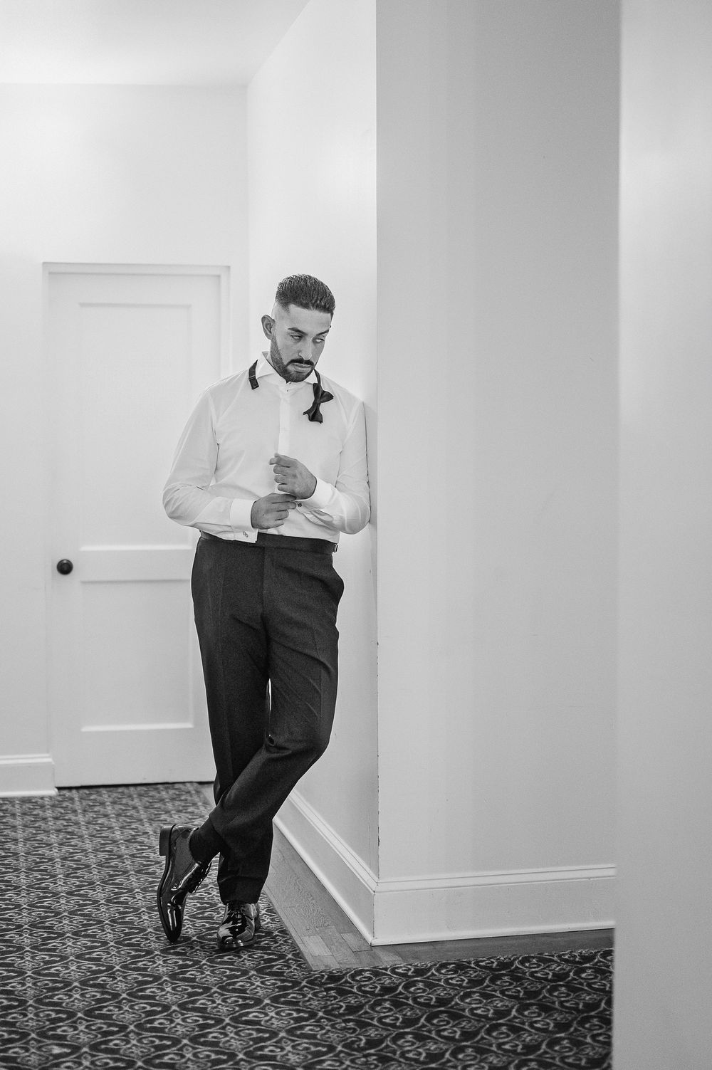 A person in formal attire leans against a white wall in a room with patterned carpet, shown in black and white.