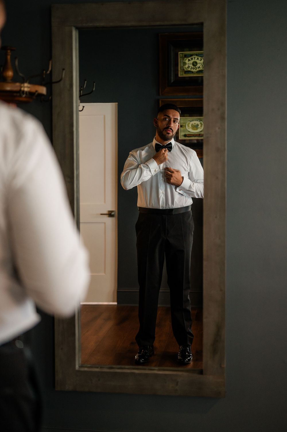 A groom adjusts his tie while getting ready in a dimly lit room with a mirror reflection.
