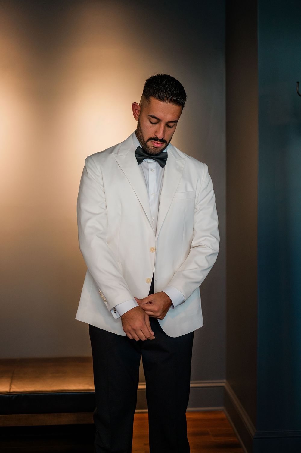 A groom in a crisp white tuxedo jacket and black pants poses thoughtfully in an elegant indoor setting.