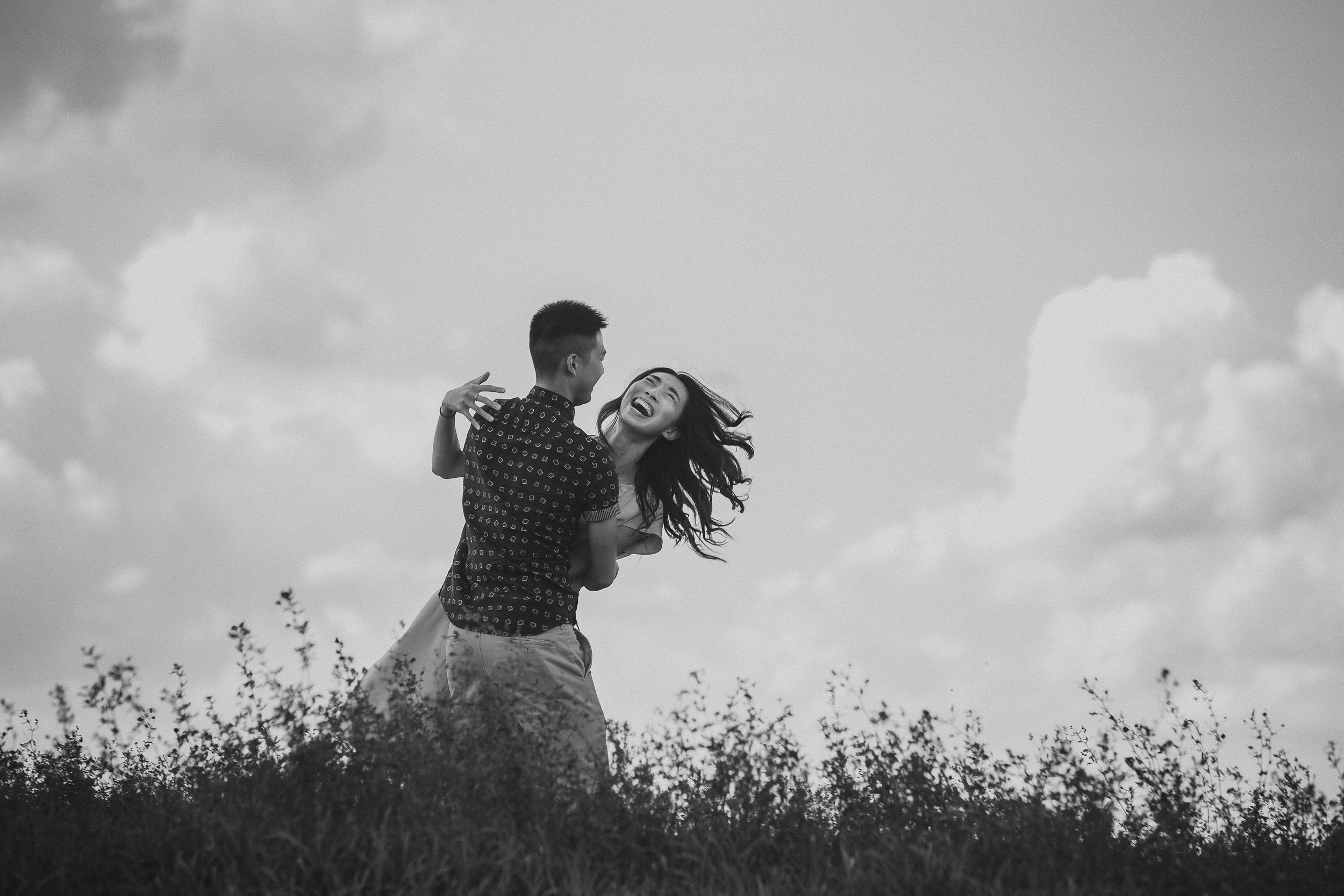 A couple dances together in a field with flowing hair against a cloudy sky in black and white.