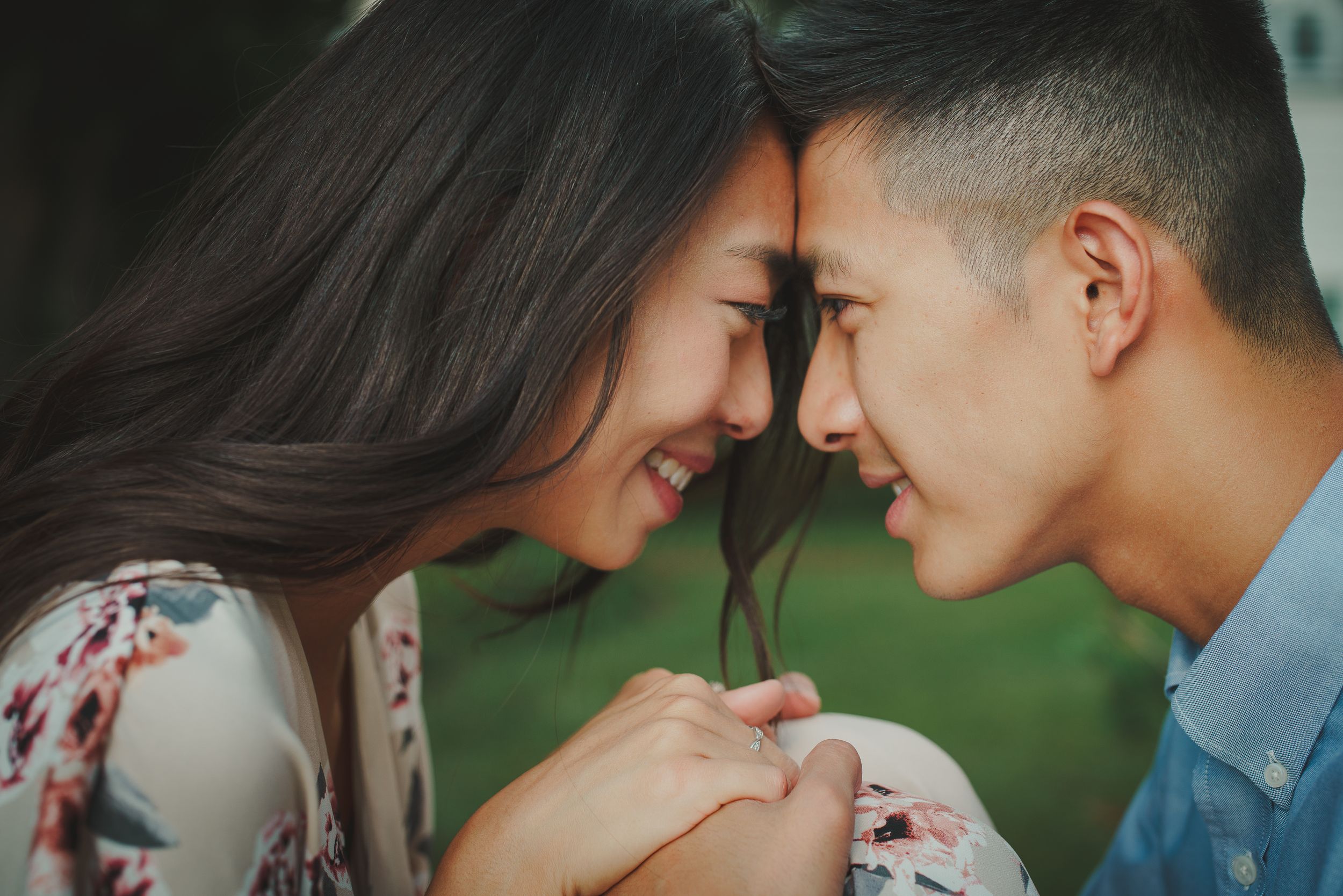 A romantic couple shares an intimate forehead touch while holding hands against a blurred green background.