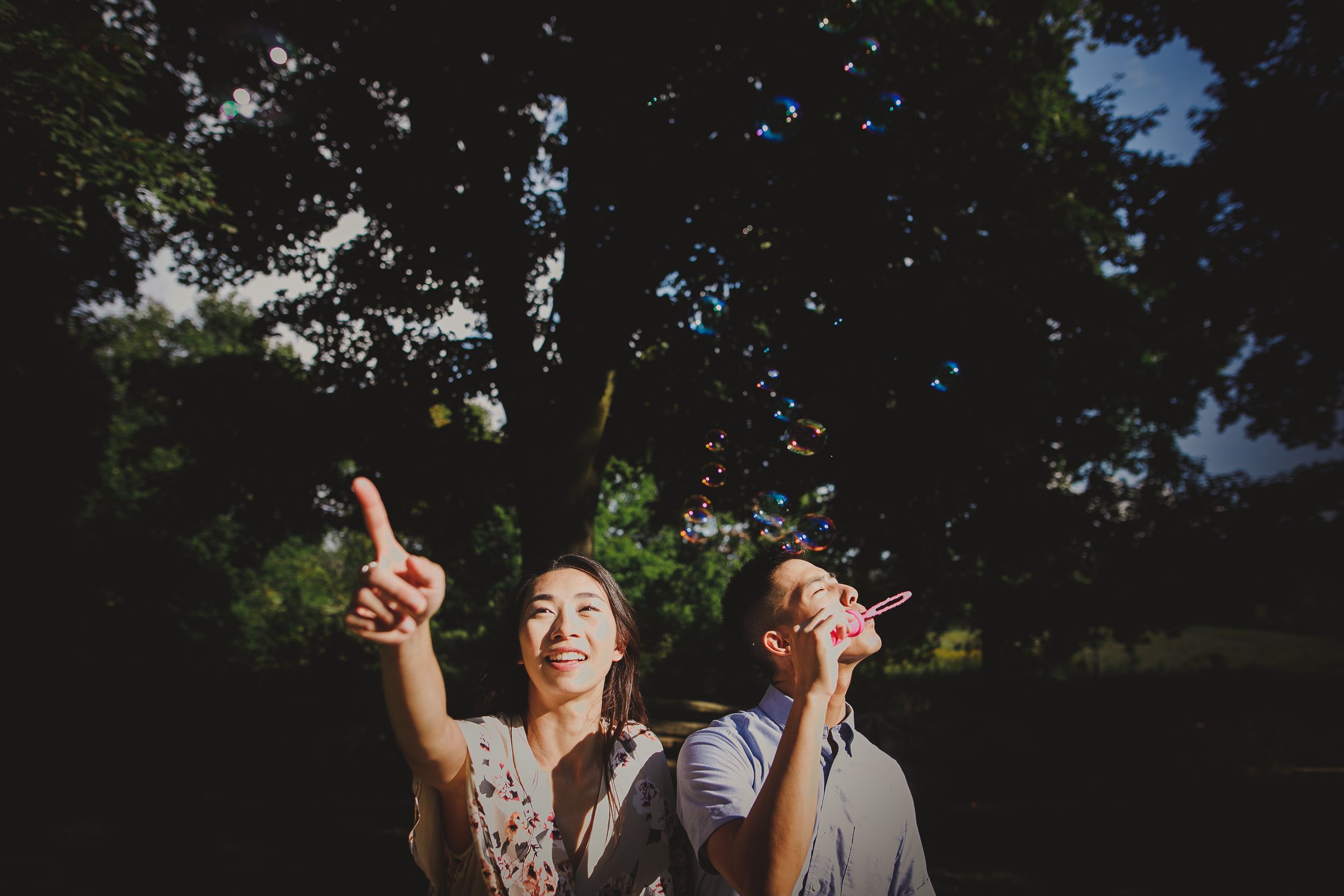 Two people point upward while playing with bubbles in a shaded outdoor setting.