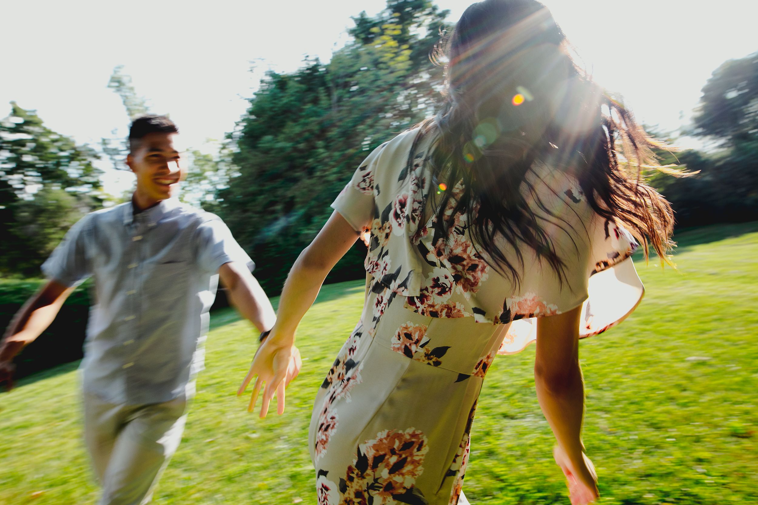 A couple runs playfully through a sunlit grassy field on a summer day.