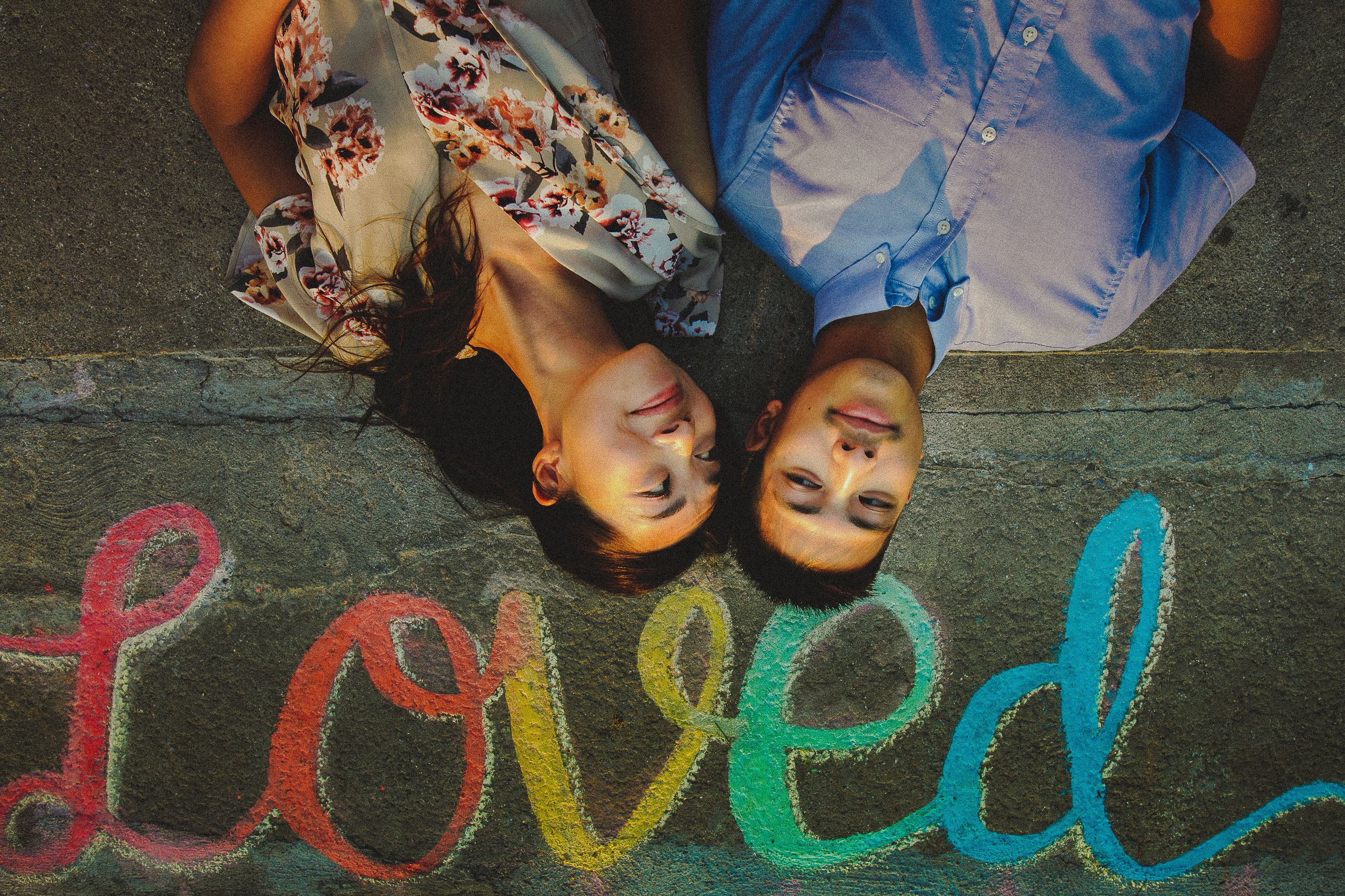 Colorful chalk writing of the word 'loved' on pavement with people's feet visible at the top of the frame.